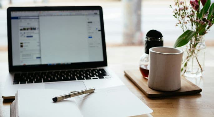 A desk with an open laptop, pen, paper, and coffe cup. 