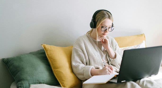 A woman seated on a couch, taking notes from a laptop.