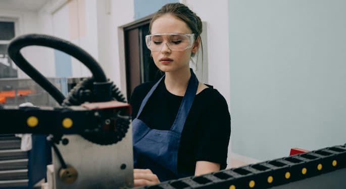 Image: A young woman in safety googles and an apron standing in front of assembly line machinery.