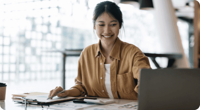 A smiling woman at a desk with a computer, calculator, and printed charts.