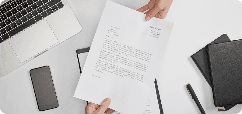 Image: an aerial view of a handoff of a grant proposal over a white desk with a laptop, phone, and two notebooks on it.