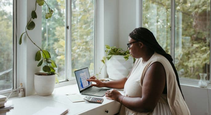 Image: A woman sitting at a white desk with lots of plants, facing windows that overlook a lot of trees. She's working in a small laptop, and has notepads and a calculator next to her.
