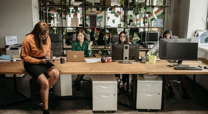 Image: four people sitting at a bank of computers at an office with lots of plants. 