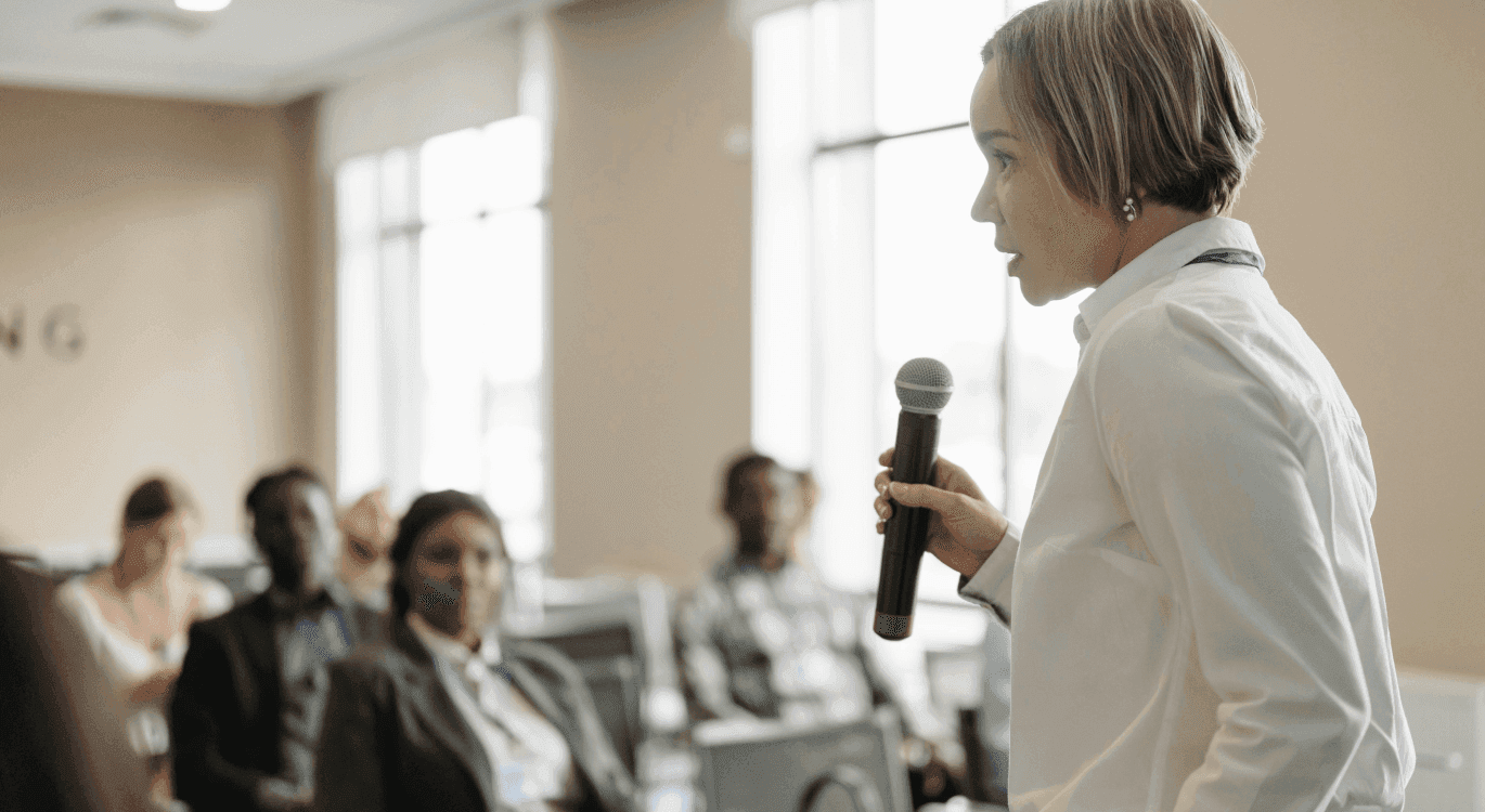 Image: a woman in front of a classroom holding a microphone and speaking to her audience.