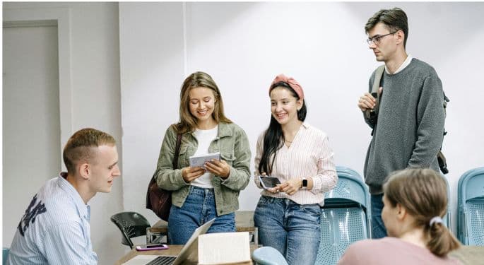 No text. Image: young coworkers or students gathered around the desk of a peer while he's on his laptop.