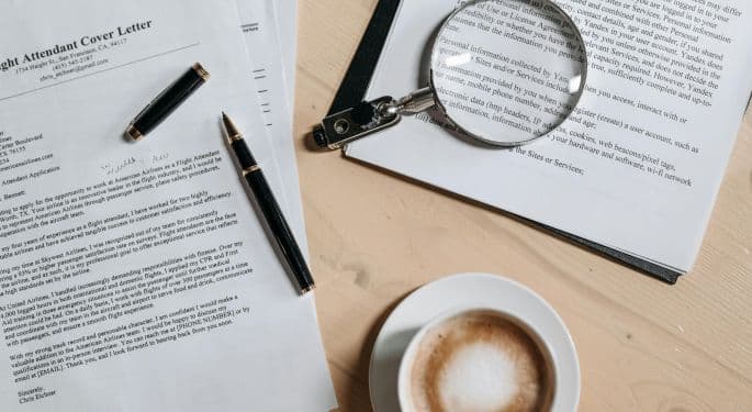 An aerial view of wooden table with several drafts of a cover letter strewn around, a magnifying glass, a fountain pen, and a latte in a white coffee cup