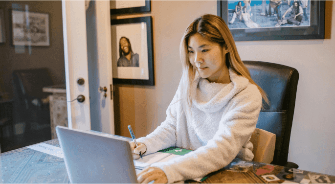 Image: A woman in a white cashmere cowl-neck sweater typing on a laptop at her home office.