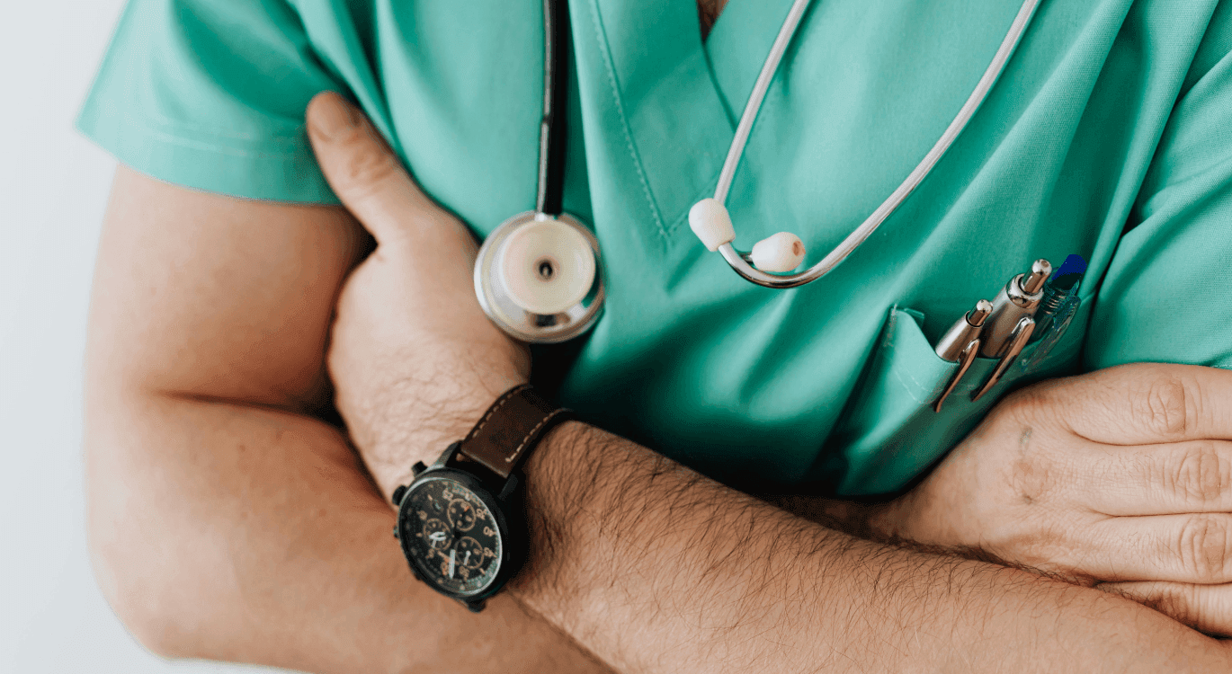 Image: a shoulder to navel shot of a male nurse in teal scrubs, black watch, wearing a stethoscope, carrying three pens in his pocket. His arms are crossed. 