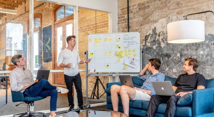 image: four men sitting in a modern office on couches while one points at a calendar 