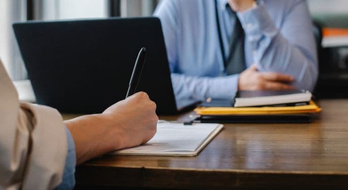 Two office workers across a desk from each other, one taking notes.