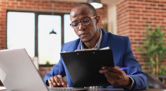 A man in a suit comparing content on a clipboard to a laptop screen.