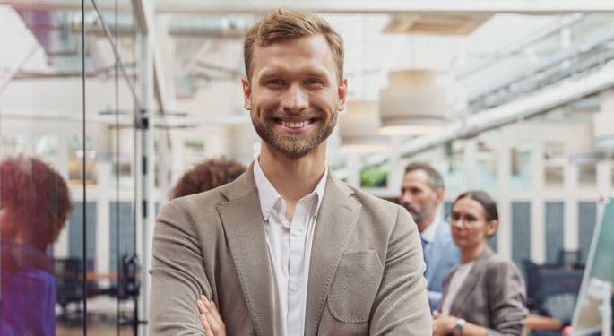 A man smiling with arms folded in a corporate workspace.