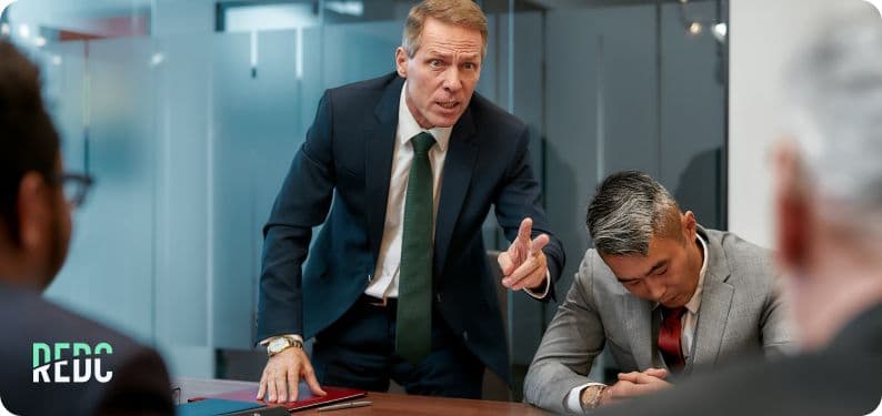 An angry-looking manager telling employees around a table what to do. 