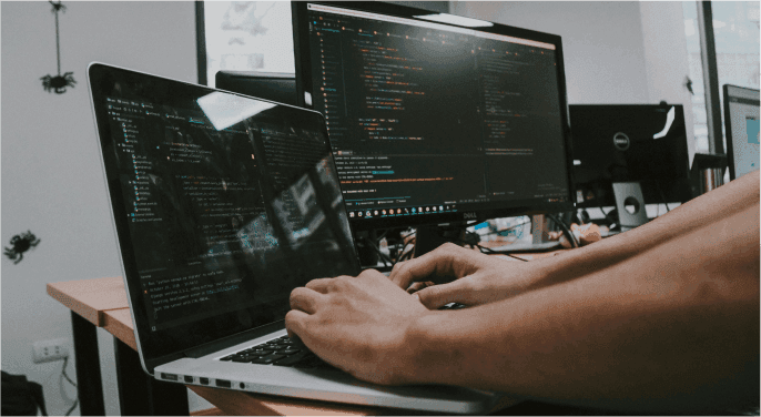 A cybersecurity professional typing on a laptop next to his dual monitor setup