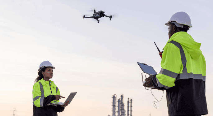 Image: Two workers, one is holding a laptop and the other is holding a drone controller. They're talking on walkie talkies and wearing visibility jackets and hard hats.